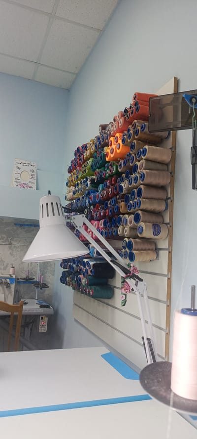 The image shows a brightly lit interior of what appears to be a tailoring or sewing workshop in Cheboksary, Russia. The wall is lined with an extensive collection of spools of thread in various colors, neatly organized on a display rack. In the foreground, a white adjustable lamp illuminates a white tabletop, which may be a workspace. To the left, a sewing machine and a wooden chair are visible, suggesting ongoing work. The room has a light blue wall and a white suspended ceiling with grid panels. A framed decorative piece with text in Cyrillic hangs on the wall. The overall impression is one of an organized and functional creative space.