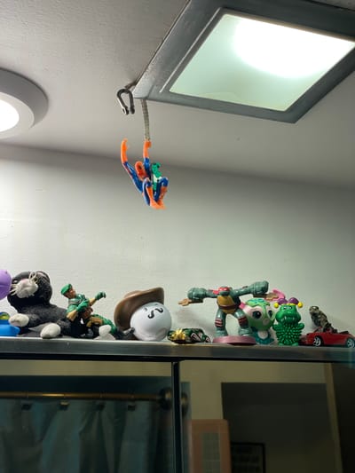 The image depicts a view of a room interior with various objects affixed to a wall or shelves. Near the top left, there is a rectangular light fixture with a bright light emanating from it. Extending from the fixture, a hook-like device is attached to what appears to be a plastic Spider-Man toy. The Spider-Man figure is posed in a web-slinging manner.

Adjacent to these elements, to the right, a collection of toys and objects are stacked vertically against what seems to be a cabinet with reflective surfaces, possibly glass doors. These objects include a red toy car, various green and other-colored figurines, and a white spherical object with cartoon-like face markings, placed atop a brown object that appears to be a cowboy hat. Below these objects is a military-themed action figure standing on top of a black and white stuffed animal.

The background is a plain white wall. The scene is indoors and appears to be in a residential setting. There is no information to confirm the location is in Clearwater, United States of America.
