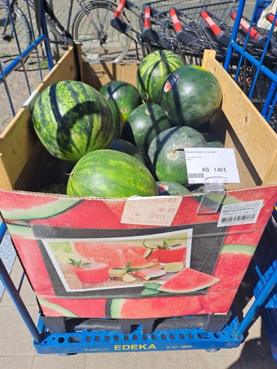 The image captures a close-up view of a wooden crate filled with watermelons, positioned on a blue plastic pallet. The crate's exterior is decorated with an illustration of watermelon slices and a glass of watermelon juice. A white price tag, clipped to the crate, indicates "Wassermelonen, rot kernarm" (red seedless watermelons), "Ursprungsland: Italien" (country of origin: Italy), and a price of "1.49 € KG". Another label below this displays "TRUDIS S.A.C." and specifies "Wassermelonen rot kernarm Klasse 3, 1,20 kg". In the background, several shopping carts with red handles are visible, as well as the wheels and frame of a bicycle. The scene appears to be outdoors, with the ground paved with light-colored tiles or concrete. The presence of shopping carts and a price tag for produce suggests a retail environment, likely a market or supermarket. The detail "E 03-2020" printed on the blue pallet suggests it may have been manufactured or last used in March 2020. The overall setting suggests a commercial area within Leipzig, Germany, where such produce and shopping amenities would be commonly found. No people are explicitly visible in the frame.