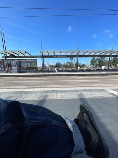 The scene is captured at the Parilly Université Hippodrome tram stop in Bron, France, under a clear blue sky. The immediate foreground shows the lower legs and feet of an individual, wearing dark blue cuffed jeans and black sneakers, situated on a grey tram platform featuring tactile paving. Multiple tram tracks run horizontally across the midground. On the opposing tram platform, a modern tram shelter with glass canopies and informational displays is visible, along with a ticket machine to the far left. A lone individual, dressed in dark outerwear and light trousers, stands near the shelter, holding a device. The background features a line of bare trees, a rack of shared bicycles or scooters, and indistinct buildings.