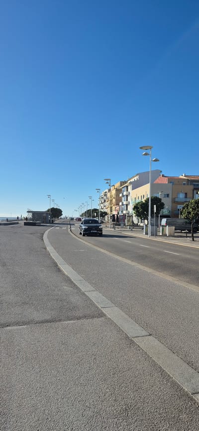 The image depicts a clear, sunny day along a coastal road in Vila do Conde, Portugal. The foreground shows a paved road with a distinct grey concrete border curving to the left. A dark-colored car is driving on the right lane of the road. On the right side of the road, there is a sidewalk lined with buildings painted in various colors, including yellow, pink, white, and blue, suggesting a commercial or residential area. Several streetlights with multiple lamps are visible along the sidewalk. In the background, a glimpse of the ocean is visible on the left, with a few people walking along a promenade or beach area. Trees are planted along the sidewalk between the buildings and the road, and there are outdoor tables and umbrellas in front of some establishments. A trash receptacle is also visible on the right side of the sidewalk. The sky is a vivid, clear blue.
