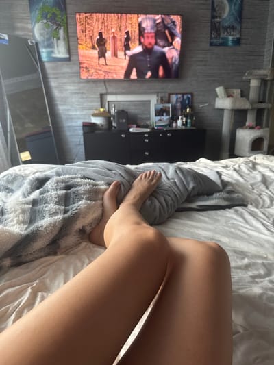 The image shows a pair of bare legs resting on a bed in a bedroom. A gray, fuzzy blanket is draped over the legs. In the background, a television is mounted on a wall decorated with textured wallpaper. The TV screen displays an image of people in a forest. To the left of the TV, a mirror reflects part of the room. A black dresser with several drawers is visible below the TV, adorned with various decorative items and bottles. On the right side of the image, a multi-tiered cat tree is situated near the wall. The overall impression is a cozy, domestic setting, possibly in Makiivka, Ukraine.