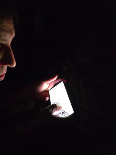 A person is holding a smartphone in their hands, illuminated by the phone's screen. The person's face is partially visible in the dark, with their nose and mouth in profile. The background is dark and indistinct, with some blurry shapes that could be plants or furniture. There is no indication of the location being Sputnik, Russia.