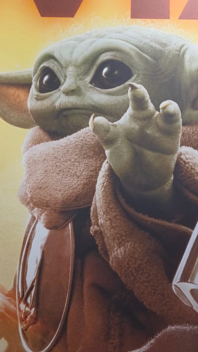This media file is a close-up portrait of Grogu, also known as The Child or Baby Yoda, from the Star Wars series "The Mandalorian." The image is oriented with a roughly 90-degree clockwise rotation, showing Grogu's head on the left and his body extending to the right.
Grogu, the main subject, has characteristic light green skin, large, dark, almond-shaped eyes with prominent reflections, and a somewhat curious or observant expression. His large ears are partially visible, and his face shows subtle wrinkles. He is enveloped in a thick, textured, brown, furry robe or blanket. One small, three-fingered hand, with short, blunt claws, is extended from the robe, appearing to reach or gesture.
In the background, a vibrant gradient transitions from bright yellow on the left to orange. On the far left, indistinct reddish-brown geometric shapes are visible, possibly part of a logo or graphic element. A metallic or silver-colored object, possibly a cup or part of his hover-pram, is partially visible in the bottom right corner, while another metallic edge is seen at the top right. The lighting is bright and warm, highlighting the textures of Grogu's skin and robe. No discernible text is visible within the frame.