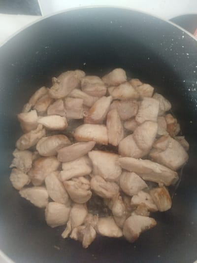 The image displays a close-up, top-down view of numerous pieces of diced, light-colored meat, likely chicken or pork, cooking in a dark-colored, possibly non-stick, pot with a speckled interior. The meat pieces are irregularly shaped and exhibit varying degrees of browning on their surfaces, indicating they are being seared or fried. A minimal amount of liquid or oil is visible at the bottom of the pot. The upper rim of the pot is visible, beyond which a blurred, light-colored surface, presumably a kitchen counter or stovetop, is present. No individuals are depicted. The scene is situated in an unspecified kitchen within the city of Klenovik, Bulgaria.