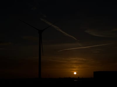 The image shows a silhouette of a wind turbine on the left side against a sunset sky. The sun is visible in the lower center, and there are contrails in the sky. The silhouette of a structure is seen on the right. The location is suggested to be Maasdijk, Netherlands, based on the prompt.