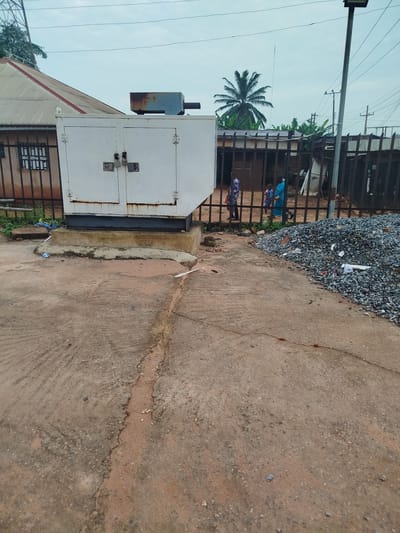 The image depicts an outdoor scene in Uselu, Nigeria, with a large white generator enclosure as the central object. The generator is positioned on a concrete base and secured with padlocks on its doors. Behind the generator, a corrugated metal roofed building with barred windows is visible. A black metal fence separates the generator area from the background, where several people, including children, are standing. Beyond the fence, lush green vegetation and a tall palm tree are prominent. A street light stands to the right of the frame, along with electrical poles and wires, suggesting an urban or semi-urban environment. A large pile of grey gravel is also present on the right side of the image. The ground is a mix of concrete and reddish-brown soil, with some cracks and debris scattered around. The sky is overcast, indicating a cloudy or potentially rainy day. The overall impression is of an industrial or service area, possibly a commercial property or a community power source, with everyday life unfolding in the background.