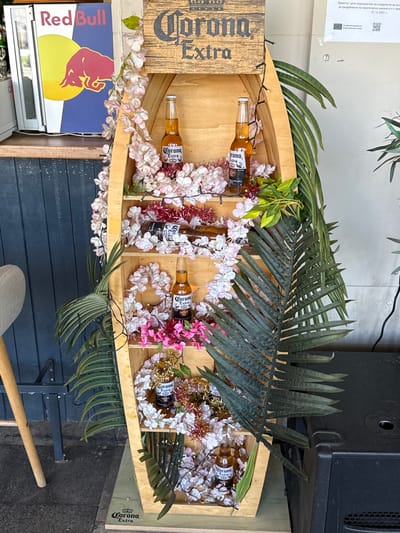 The image shows a display unit shaped like a boat, filled with decorative flowers, lights, and bottles of Corona Extra beer. A Red Bull refrigerator is visible to the left of the display. The display is situated outdoors, possibly in front of a commercial establishment, with a dark blue wooden wall in the background. A black speaker is positioned to the right of the display unit. The overall scene appears to be a festive or promotional setup. The image was taken in Vidin, Bulgaria.