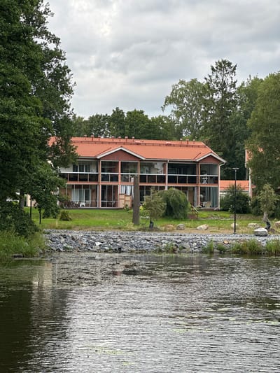The image shows a multi-story building with a red tiled roof and large windows, set against a backdrop of trees. The building appears to be residential, possibly apartments or townhouses, with balconies on at least two levels. The foreground features a body of water with ripples and some floating debris. A rocky embankment separates the water from the grassy area in front of the building. The sky is overcast. The scene is likely located in Vammala, Finland, given the context. There are no visible people in the image.