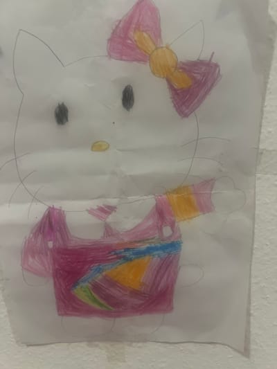 A child's drawing of Hello Kitty is taped to a textured wall. The drawing is on white paper and depicts Hello Kitty facing forward with a large pink bow on her head. She is wearing a pink dress with colorful accents and has a white hand visible on the right side of the drawing. The drawing is creased in several places. The wall appears to be painted a light, neutral color. No specific location in St. Gallen, Switzerland is discernible from the image.
