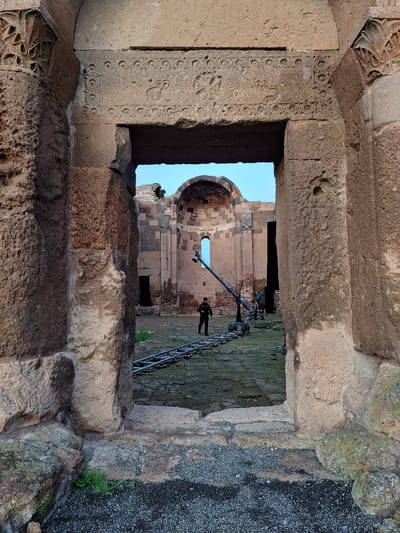 A low-angle, medium shot presents an architectural view through a stone-framed opening of an ancient structure, likely a church or similar building, located in Anipemza, Armenia. The viewer is looking through the stone opening towards the ruins. The frame's stonework on the left side is adorned with detailed carvings, with additional stone textures visible throughout the frame.
Outside the opening, a ladder and what appears to be film or recording equipment, as well as a human figure in dark attire, are visible. The ground is covered with a mixture of grass, gravel, and other vegetation. The background reveals the clear blue sky.