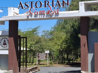 The image shows the entrance to the Astorian Nomaya, located at coordinates -0.766, 36.433 in Karagita, Kenya. The entrance is marked by a white archway with a sign reading "ASTORIAN Nomaya" in red lettering. The archway is supported by two large brown pillars and a metal gate. Behind the archway, there is a dirt path that leads into a grassy area. Several green trees are visible in the background, and there is a small sign on a pole that lists the services offered by the facility. The services include accommodation, holiday residency, conferencing, events/grounds, and camping. A wooden ladder lies on the ground to the left of the gate.  The image appears to be taken during the day, as the sky is bright blue and the sun is shining.