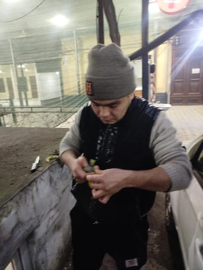 The image captures a person, likely male, in what appears to be an outdoor or semi-outdoor setting at night. He is positioned slightly off-center to the right, looking down intently at an object in his hands. He is wearing a grey knitted beanie with a distinct orange square patch featuring an "H" in white on the front. Below the beanie, he has on a light grey long-sleeved shirt, over which he wears a dark, possibly black, sleeveless vest. The vest has a patterned or textured detail on the chest area.
The main activity shown is the person actively peeling a yellow, citrus-like fruit, possibly a lemon or lime. His hands are engaged in the task, and he holds a small tool or another knife to assist with the peeling. On a rough, light-colored concrete or stone ledge to his left, a small, silver-bladed knife with a white handle is resting.
The background is somewhat blurred due to the shallow depth of field and low light conditions. To the left, there's a light-colored building or structure visible through what appears to be a fine wire mesh or netting. This area is dimly lit. To the right, behind the person, there's a brightly lit area featuring a dark wooden double door or entrance, and a bright light source, possibly a streetlamp or a sign, casting a strong glow. A small, white object, possibly a sink or basin, is also faintly visible near the door. On the far right, the side of a white vehicle, likely a car, is partially visible. The overall lighting suggests it is nighttime or very late evening, with various artificial light sources illuminating the scene. The atmosphere is casual, and the person's expression is one of focus on the task at hand.