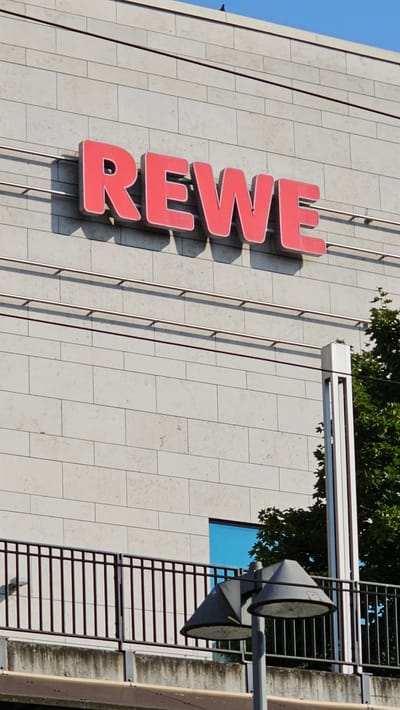 A sign with the word "REWE" in red capital letters is mounted on the exterior wall of a building. The wall is made of light-colored rectangular blocks. Below the sign are two horizontal metal rails. Further down, there is a black metal fence. In front of the fence are two street lamps with conical shades. To the right of the fence, a section of a tree with green leaves is visible. A bright blue window can be seen behind the fence. The sky above is clear and blue. The scene is located on a sunny day in Neu-Ulm, Germany.