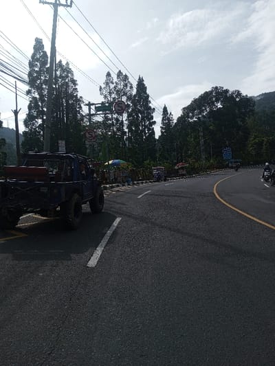 The image depicts a roadside scene on a clear day, with an asphalt road curving to the right. In the foreground, a dark blue off-road vehicle, possibly a modified Suzuki Jimny with large knobby tires and an external roll cage, is positioned on the left side of the road, with an occupant visible. White and yellow road markings are present. A tall utility pole supports multiple overhead wires on the left. Above the road, a green sign displays white text in Indonesian, including "Jl. Raya Puncak," suggesting a location in Indonesia. Two red circular "No Stopping" signs are also visible. Along the right side of the road, behind black and white striped barriers, are several vendor stalls, one featuring a multi-colored umbrella. People are visible near these stalls and riding motorcycles further down the road. Dense vegetation, including tall coniferous trees and a distant forested mountain, frames the scene under a sky with scattered white clouds. The location is specified as Sandamal Eliya, Sri Lanka.