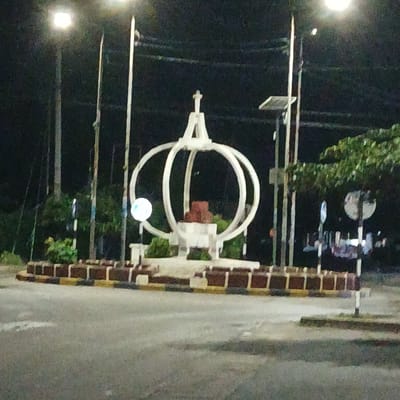 The image displays a nighttime scene of the Tanga Clock Tower Roundabout, also known as the Uhuru Roundabout, located in Tanga, Tanzania. At the center of the roundabout is a prominent, white, crown-shaped monument. This monument is constructed from multiple intersecting white arches, forming a large, open spherical cage structure, topped with a distinct finial that appears pointed or cross-shaped. Within the monument's base, a reddish-brown, multi-tiered block structure is visible, encircled by green low-lying foliage.
The monument rests on a raised circular island, which is bordered by a low curb composed of alternating dark reddish-brown and light yellow/white rectangular blocks. The foreground depicts a dark asphalt road surface. The area is illuminated by several bright overhead streetlights mounted on tall poles. One pole on the right side of the frame features a small solar panel, while another pole to the left of the monument displays a spherical, glowing blue-white light fixture. Numerous other slender poles, consistent with utility or signage infrastructure, are present around the roundabout. Dark green foliage and trees are visible in the background against the dark night sky. No individuals or vehicles are discernible within the frame.