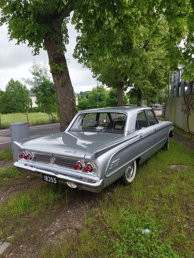 A silver/grey vintage four-door Mercury Comet sedan is parked on a grassy verge next to a road in Manternach, Luxembourg. The car, a prominent subject, displays classic styling with abundant chrome trim, including bumpers, window surrounds, and a distinctive rear panel. It features a chrome grille, stacked triple red taillights, and three chrome vents on the rear quarter panel. White-wall tires are visible. The rear license plate clearly reads "18355" and includes a small "L" for Luxembourg, along with a white circular sticker or marker with the number "7".

The setting is an outdoor, verdant scene under an overcast sky. A large tree with lush green foliage dominates the left side of the frame, its trunk and canopy extending across much of the background. Additional trees and greenery are visible further back, hinting at a rural or suburban landscape with distant hills or structures. Behind the front of the car, a metal fence and a wall with abstract art are present. The ground is a mix of green grass, clover, and some areas of dirt or mud. A small, dark utility box sits near the car's rear. No people are visible, and the car appears to be stationary, presented in a calm, static pose.
