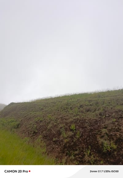 The image shows a hillside covered in grass and sparse vegetation, with a dense fog obscuring the background and sky. The foreground features a grassy slope on the left, transitioning to a more rugged, earthy terrain on the right. The top of the frame is a solid expanse of white, indicating the thick fog. Text overlays at the bottom of the image read "CAMON 20 Pro" and "24mm f/1.7 1/201s ISO50". The overall impression is a natural, outdoor scene in foggy conditions. The location is identified as Nguroje, Nigeria.