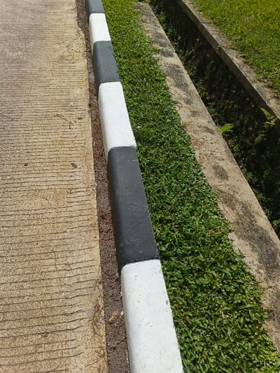 A concrete sidewalk with parallel grooves meets a curb painted with alternating black and white stripes. Between the curb and a concrete drainage ditch grows dense green grass. More grass is visible beyond the drainage ditch. The concrete surfaces are weathered and textured. The scene is bathed in daylight.
