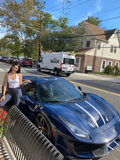 The image displays a young adult female posing next to a dark blue Ferrari 488 Pista on a sunny day. The female, with long dark hair, wears a white halter top and dark grey leggings, smiling directly at the camera while holding keys in her right hand. The Ferrari, featuring triple white racing stripes on its hood and yellow brake calipers, is parked on the right side of a paved street with a solid yellow center line. In the background, a white cargo van belonging to "DREWING PLUMBING" (contact information "201.298.5080 | drewing.com") is visible on the street, equipped with a roof rack. Beyond the van, a multi-story commercial building with light-colored siding, a brown brick lower facade, and a brown shingled roof is present; a "Keller Williams" sign is affixed to its facade. Utility poles, power lines, and deciduous trees with partially browned foliage line the street. Additional vehicles are visible in the distance. The scene is located on a street in Ridgewood, New Jersey, United States, specifically identifiable as the vicinity of 257 East Ridgewood Avenue, where the Keller Williams office is situated.