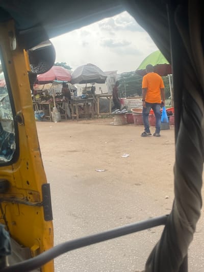 The image depicts an outdoor scene, likely a marketplace, in Galadimawa Village, Nigeria. In the foreground, a yellow vehicle, possibly a rickshaw or a similar motorized tricycle, is partially visible. The perspective suggests the image is taken from inside this vehicle. In the mid-ground, a market area is set up with wooden stalls and various goods displayed. Several individuals are present, some tending to the stalls and others browsing. One man, wearing an orange t-shirt and blue jeans, is walking away from the viewer, carrying a blue plastic bag. Umbrellas, one green and maroon, and another pink, are erected to provide shade. Buildings, seemingly residential, are visible in the background. The ground is paved with dirt and asphalt. The overall atmosphere suggests a typical day at a local Nigerian market.