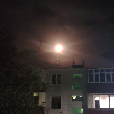The image depicts a nighttime scene in Bukhara, Uzbekistan. The sky is dark and cloudy, with a bright, luminous full moon dominating the upper center of the frame. The moon appears to have a halo or glow around it, possibly due to atmospheric conditions or the camera's exposure.
In the foreground, a portion of a leafy tree is visible on the left side, its dark silhouette contrasting with the bright sky. Below the moon and tree, the upper floors of a building are visible. The building is light-colored and appears to be an apartment complex. Several windows are visible; some are dark, while others are illuminated with a greenish or yellowish light, suggesting interior lighting. One window on the right side of the building is particularly bright, with a curved arch detail above it.
Two dark, slender antenna-like structures rise from the roof of the building, barely discernible against the dark sky. The overall mood of the image is quiet and somewhat mysterious, with the bright moon creating a dramatic focal point against the darkness and the architectural elements of the building. The time of day is clearly night, and the weather appears overcast, with the moon shining through the clouds. There is no visible text in the image.