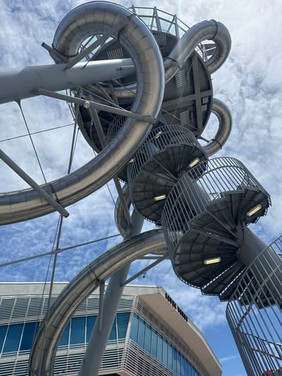 The image shows a low-angle, wide shot of a large, outdoor spiral slide structure in Aventura, Florida. The slide, made of shiny, segmented metal tubing, curves and twists upwards against a partly cloudy blue sky. Several grey, metal spiral staircases with railings are visible, attached to the slide's support structure. The staircases are illuminated by fluorescent lights. In the background, a modern building with blue windows and horizontal silver louvers is partially visible. The building has signage that reads "LEVEL THREE". The overall impression is of a large, complex, and modern amusement or attraction.