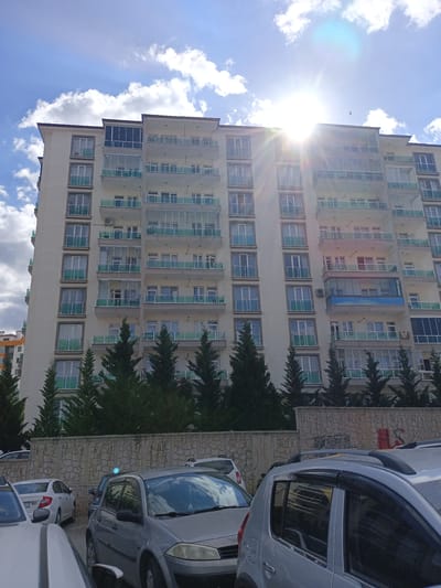 The image shows a multi-story apartment building in Yahşihan, Turkey, under a partly cloudy sky with the sun prominently visible and causing lens flare. The building has a light-colored facade with numerous windows and balconies, many of which are enclosed with glass. Green railings are visible on some of the balconies. In front of the building, there are several evergreen trees and a stone wall with a textured pattern. Below the wall, a parking lot is visible with several cars parked, including a silver sedan and a gray hatchback. The cars appear to be typical civilian vehicles. The overall scene suggests a residential area in a city environment. There are no people visible in the image.
