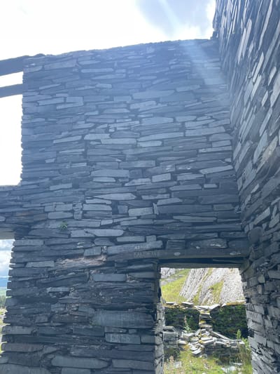 The image depicts the interior of a derelict stone structure, likely a ruin, constructed from dark, flat slate stones in Tanygrisiau, United Kingdom. The walls are unevenly stacked, with visible mortar and gaps between the stones. Sunlight streams in from the upper left, casting lens flares and illuminating the rough texture of the stonework.

Several graffiti markings are visible on the walls. In the center of the main wall, above a lower opening, are the words "ЖАННА" and "КАДЯИСЛАВ" written in capital letters. Below this, another opening reveals a view of a green, rocky landscape with scattered debris and vegetation. Further down the main wall, the word "BRED" is etched. On the right wall, also partially visible, is writing that appears to read "PA".

The structure is open to the elements, with portions of the roof and ceiling missing. The ground inside the ruin is covered in loose stones and some patches of grass. The overall impression is one of decay and abandonment in a natural, rugged setting.