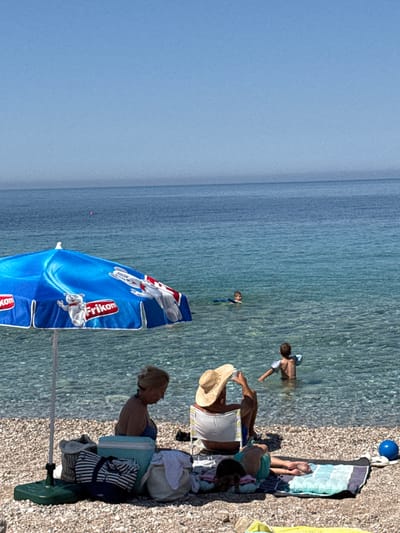 In Perazića Do, Montenegro, a sunny beach scene unfolds. A bright blue umbrella, adorned with white cartoon bears and the "Frikorn" logo, stands prominently on a pebbled shore. Underneath it, a woman with blonde hair tied back in a bun sits, looking down, while near her are several beach bags and a cooler. Further along the beach, another individual wears a wide-brimmed straw hat and is seated in a beach chair, facing the sea. To the right, a child lies on a striped towel, their back to the camera. In the crystal-clear water, two young boys are visible, one in the middle distance with an arm float, and another closer to the shore, also with arm floats. The calm, blue sea stretches to the horizon under a clear sky.