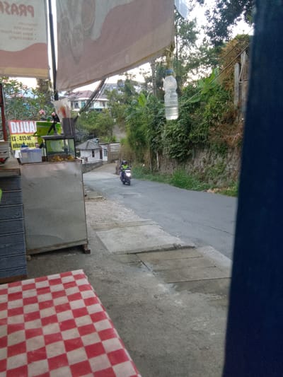 This media file captures a street scene in Cisarua, Indonesia, likely during daylight hours given the ambient light. The foreground is dominated by a table covered with a red and white checkered tablecloth, suggesting an outdoor seating area for a food stall or restaurant.
To the left, a silver food cart with a glass display case is visible, containing some fruit, possibly oranges, and other items. A sign above the cart reads "DIJUAL" in large red letters, meaning "FOR SALE" in Indonesian. Below this, "LT: 415 M²" indicates the land area is 415 square meters, and "HUBUNGI: 0819" is a contact number fragment. Behind the cart, a structure with a white exterior and a red-tiled roof can be seen, typical of Indonesian residential architecture.
A motorcycle rider, wearing a helmet and a green jacket, is traveling down the asphalt road. The road is flanked by a stone wall covered in greenery and trees, forming a natural embankment. A plastic bottle of water is hanging from a wire, possibly for shade or decoration. The overall impression is one of a casual, everyday scene in a semi-rural or suburban area. The time of day appears to be late afternoon or early evening, with the lighting being somewhat diffused.