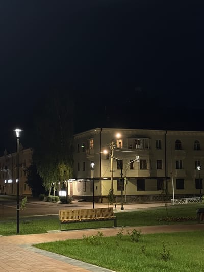 This is a night-time, slightly elevated, full-frame shot of a paved walkway with a grassy area and a wooden bench in the foreground. The walkway has a reddish-brown brick pattern with lighter colored edging. To the right of the walkway is a patch of well-maintained grass, dotted with small green bushes and saplings. A long, light-colored wooden bench with a slatted back and seat is positioned in the grassy area, facing towards the left of the frame.
Beyond the grassy area, a street runs horizontally across the mid-ground. On the other side of the street, a building complex stretches across the frame. The building is light-colored, possibly beige or cream, and appears to be several stories tall with many windows. Some windows are illuminated from within, suggesting occupancy. There are balconies and architectural details on the facade. Streetlights are visible along the street, casting pools of light. One prominent streetlight with a modern design stands to the left of the frame. Trees, including a large, leafy one, are situated between the street and the building. The sky above is dark, indicating nighttime. The scene depicts a quiet urban environment, likely a park or public space adjacent to residential or commercial buildings in the city of Radkevshchina, Russia.