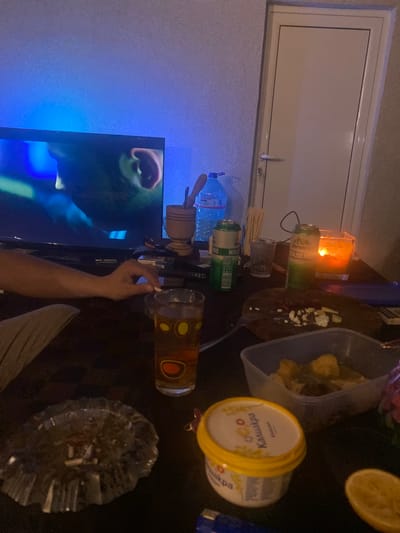 This image captures an indoor, dimly lit evening scene, likely a casual gathering or an individual relaxing. The perspective is from above a table, which is cluttered with various items. A person's arm and hand are visible in the lower right, seemingly reaching for something or holding a small item near a patterned glass containing a brown liquid, likely beer. An ash tray filled with cigarette butts and ash sits to the right, suggesting smoking.
On the table, several beverages and snacks are arranged. There's a blue plastic bottle labeled "DEVIN" (water), two green cans (one identifiable as "Пиринско" beer, another with "МОР"), and a yellow-lidded container marked "Кауфланд" (likely a dairy product like spread cheese or yogurt). A clear plastic container holds what appears to be fried potatoes or other snack food, accompanied by a fork. Half a lemon slice is also on the table. A lit candle in a glass holder provides warm illumination, contrasting with the cool blue light emanating from a television screen on the left, which displays a close-up of a person's face or head.
Other items include a wooden mortar and pestle, a small clear glass, and a black remote control. A white-framed window with a light-colored blind or panel, displaying "LED" on its handle, is visible in the background. The visible text on the items, such as "Пиринско" and "Кауфланд," are consistent with the provided location context of Belopoptsi, Bulgaria. The overall atmosphere suggests a relaxed, low-key indoor activity.