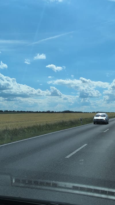 An outdoor, eye-level, medium shot captures a car driving on a paved road in Buttstädt, Germany. The car, a silver four-door sedan, is traveling in the right lane towards the viewer. The road is lined with a grassy shoulder and a field of golden wheat. In the background, a line of trees and rolling hills can be seen under a bright blue sky dotted with white clouds. The overall scene depicts a rural landscape under a sunny sky.
