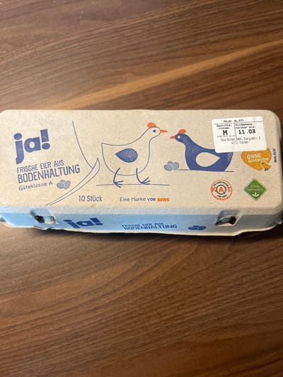 The image shows a cardboard egg carton with 10 eggs. It is laying on a wooden surface. The carton is blue and white with the word "ja!" written on it. The carton has the words "frische Eier aus Bodenhaltung," which translates to "fresh eggs from free-range." The carton also has a sticker on it that says "M 11.03," which is a date.  The coordinates provided place this scene in Köln, Germany.