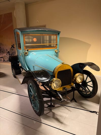 A vintage, light blue automobile, likely from the early 20th century, is displayed indoors in what appears to be a museum or exhibition space in Wassenaar, Netherlands. The car features a prominent brass radiator grille and two large, circular brass headlights. Its body is painted a distinctive light blue, complemented by black fenders and black-spoked wheels with light blue hubs and pneumatic tires. The cabin showcases intricate wooden window frames, and a small, circular orange or pink object is visible through a rear window. A black step or running board is attached to the side.
The background consists of plain light beige walls, with a large, white cylindrical column visible on the left side. Black ropes act as barriers in the foreground and to the right, reinforcing the exhibition setting. Shadows are cast on the walls and the light-colored tiled floor, indicating artificial lighting. A partially visible framed poster on the lower left wall displays the text "LON AUTOMOBILE," strongly suggesting "SALON AUTOMOBILE," a term commonly used for motor shows. The car is stationary, presented as an exhibit.