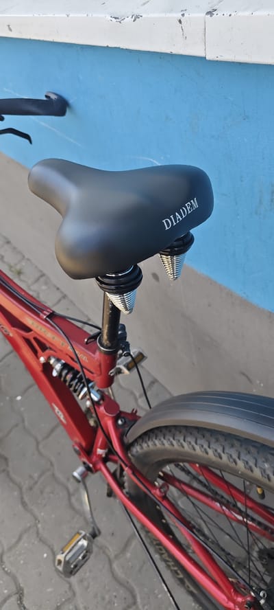 A red full-suspension bicycle is parked outdoors, leaning against a blue wall. The bicycle seat, black with the word "DIADEM" embossed in white, is clearly visible, supported by two coiled silver springs. The frame of the bicycle is red, and a portion of the rear tire and fender is also visible. The ground is paved with interlocking grey bricks. The scene appears to be in a city or town, with the blue wall suggesting an urban setting. No people are present in the image. The lighting suggests it is daytime, possibly late afternoon, given the shadows. The location is stated to be the city of Mezhdurechensk, Russia.