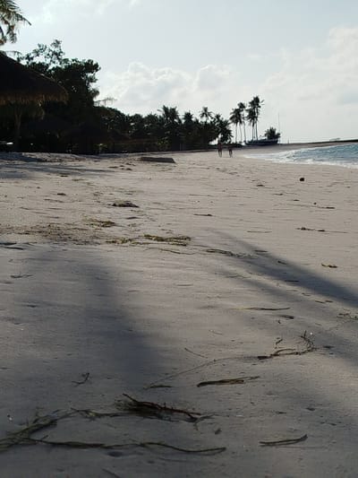 The image depicts a sandy beach in Hinnavaru, Maldives, with the ocean and palm trees in the background. The foreground is dominated by the beach, with some seaweed scattered across the sand. Two figures are walking in the distance along the shoreline, their backs to the camera. A thatched-roof structure is visible on the left side of the image, partially obscured by trees. The sky is partly cloudy, and the sun casts shadows across the sand.