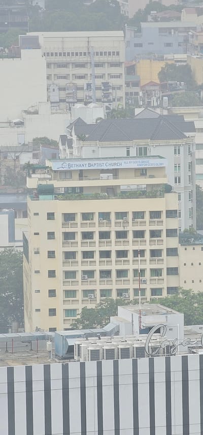 The aerial view captures a section of the Makati City skyline, Philippines, on a hazy day. The prominent feature is a beige, multi-story building with a repeating pattern of windows and visible air conditioning units. Above this building, a banner displays "BETHANY BAPTIST CHURCH" with the text "ALL BY GOD'S GRACE AND FOR GOD'S GLORY" to its right. Behind this, a building with a dark, pitched roof is partially visible, and further in the background, a cluster of buildings in varying shades of white and grey, interspersed with greenery, suggest a dense urban environment. In the foreground, a section of a building with a distinctive pattern of alternating black and white vertical stripes is seen. The overall scene is dominated by architecture and lacks any discernible human presence or activity.