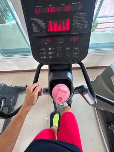 The image shows a first-person perspective of someone using an elliptical machine in what appears to be an indoor gym setting. The person is wearing bright red cycling shorts and neon yellow socks. Their hands are visible on the machine's handlebars. A pink water bottle is placed in a holder on the console.
The console of the elliptical machine displays several metrics. The time is 8:25, and the resistance is 8.88. The distance is 5.1, and the wattage is 80.0. Calories burned are shown as 289.8. A heart rate zone graph is also visible on the console, along with buttons for various functions like "Quick Start," "Enter," "Target," and "Stop." Some text on the console provides a safety caution about consulting a physician before starting a fitness program and reading the equipment's manual.
The background suggests an indoor environment with a window offering a view of greenery, hinting at daylight. The floor is tiled. The overall scene depicts a fitness activity in progress.