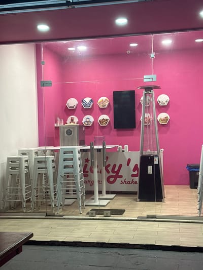 This image captures the interior of a brightly lit establishment, possibly a cafe or dessert shop, viewed from outside through a glass facade. The primary color of the interior walls is a vibrant magenta pink. The floor is light-colored tiling.

No people are visible in the scene. The activity suggested is that the business is either closed or preparing to close, indicated by several white bar stools stacked neatly on top of each other, pushed against white tables or counters.

On the pink wall, a black flat-screen television is mounted. Below and to its right, there is a large, tall, silver and black outdoor patio heater. Also affixed to the pink wall are several hexagonal and circular display shelves, each holding what appear to be various food items or dessert packaging. A white counter below the TV also holds some equipment, possibly for preparing drinks. A black trash can is visible in the upper right corner.

Visible text on a white counter or sign in the middle right of the image clearly reads "Lucky's shake" with the word "burger" partially visible underneath, suggesting the establishment might offer both. The overall lighting is artificial, with oval-shaped lights on the left wall reflecting off the glass, implying it could be evening or night. The scene depicts a clean and organized interior, located in Egkomi Lefkosias, Cyprus.