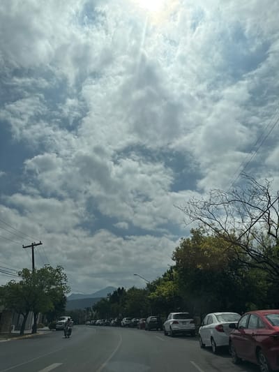 The image captures a street scene in San Pedro Garza García, Mexico, on a day with scattered clouds. The road curves slightly, with parked cars lining the right side. A group of motorcyclists is visible on the road. Power lines stretch across the sky. Trees are abundant, lining the street. A mountain is visible in the distance, partially obscured by the clouds.