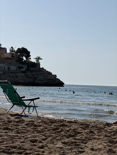 This image depicts a beach scene in Finestrat, Spain. In the foreground, a green beach chair is positioned on the sandy shore, facing the water. A blue towel is placed beneath the chair. The water is calm with small waves breaking near the shore, reflecting the sunlight. Several people are visible swimming and wading in the sea. In the background, a rocky cliffside rises from the sea, topped with vegetation and buildings. The sky is clear and blue. The overall environment suggests a sunny day at the beach.