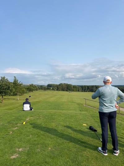 The image shows a golf course in Pocklington, United Kingdom, at the coordinates 53.939, -0.757. The scene is on the green, with two golfers on the tee box. One golfer is sitting down, facing away from the camera, and holding a golf club. The other golfer is standing, facing the camera, holding a golf club and wearing a white cap and a light blue sweatshirt. The golfers are facing a golf course that extends towards the horizon, where a group of golfers are in the distance. The course is green, with a forest line beyond it. The sky is blue and clear, with a few white clouds. The ground is covered in lush grass. The golfers are about to play a round of golf.