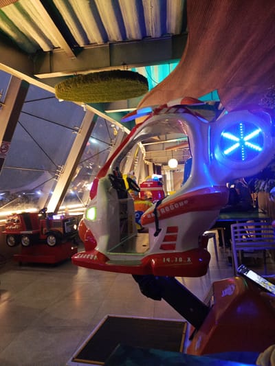 The image depicts a children's amusement ride designed to resemble a helicopter, with red and white detailing and blue LED lights on its rotor. The ride is positioned within an indoor entertainment venue. To the left, a red toy truck ride is visible. The environment features a ceiling with exposed beams and a textured surface, along with large windows or glass partitions. The flooring appears to be tiled. In the background, other rides and seating areas can be seen, suggesting a family entertainment center. The overall lighting is dim, with the LED lights of the rides being prominent. The helicopter ride features text that reads "Sky Rescue" and "14.70.X". This scene is located in the city of Guayaquil, Ecuador.