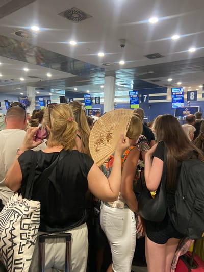 The image depicts a queue of people at what appears to be an airport or a transportation hub, specifically within Mrčevac, Montenegro, likely near Tivat Airport given the context of flights. The scene is indoors, characterized by a brightly lit ceiling with multiple light fixtures and air vents.
In the foreground, a woman with blonde hair tied in a ponytail is holding a pink mobile phone in her right hand, seemingly taking a photo or video. She is wearing a black top and is standing behind a large patterned tote bag and a suitcase handle. Next to her, a person holding a decorative fan with an ornate pattern is partially visible.
Further back, the queue continues with individuals facing forward. Several digital display screens are mounted on the blue and white walls in the background, showing flight information. One screen displays "FLYONE 5F6822 Chisinau" with a time of "14:05". Other screens indicate gate numbers, such as "7" and "8".
The overall atmosphere suggests a busy travel environment, with people waiting their turn. The lighting is bright and consistent with an indoor public space. The environment is clean and organized, with visible infrastructure related to transportation services.