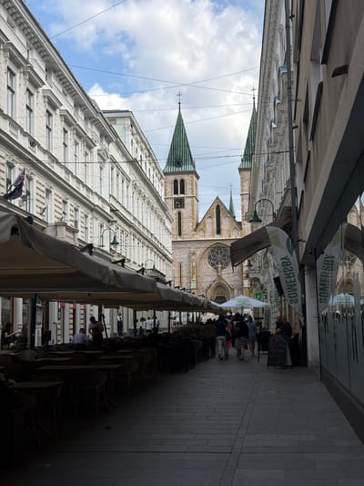 The image depicts a street scene in Sarajevo, Bosnia and Herzegovina, likely in the historic center. On the left, a long, multi-story building with ornate white facade and numerous windows lines the street. Overhanging awnings of a cafe extend across the sidewalk, shading rows of tables and chairs, some of which are occupied by people. In the background, a prominent stone cathedral with twin green spires and a large rose window is visible. The street is paved with stone and a number of people are walking along it. On the right side of the street, another building with a plain facade and windows is present, with advertisements and signage visible. The sky above is partly cloudy.