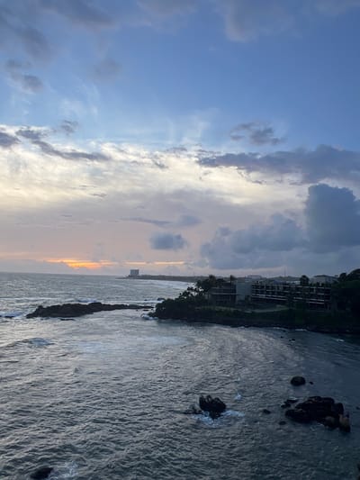 The image depicts a serene coastal scene in Galle, Sri Lanka, likely captured during sunset. The sky is a dramatic canvas of blues, grays, and soft oranges, with clouds stretching across the frame, hinting at the transition from day to night. The ocean, rendered in deep blues and grays, is calm, with gentle waves lapping at the shore and around dark, jagged rocks that jut out from the water.
Along the coastline, a cluster of modern buildings, likely hotels or apartments, are visible, nestled amongst lush greenery. Their dark facades stand in contrast to the softer hues of the sky and sea, suggesting an urban development integrated with the natural landscape. A distant structure, possibly a tower or lighthouse, pierces the horizon, adding a point of interest to the expansive view.
The overall mood is tranquil and contemplative, evoking a sense of peace and the beauty of nature. The soft light of the setting sun casts a warm glow, emphasizing the textures of the water and the dramatic cloud formations. The image captures a moment of natural beauty and quietude, characteristic of a coastal sunset. No text is visible in the image.
