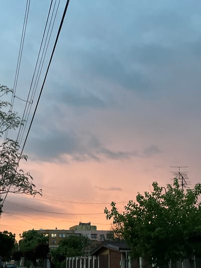 The image depicts a dusk scene in an urban setting. In the upper-right quadrant, there's a building and tree. An antenna is present on the roof of the building. In the foreground, there are wires and trees. The sky is colored with a gradient from blue to orange. The location is likely Nova Zagora, Bulgaria.