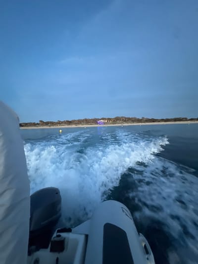 The image shows a view from a boat moving along the coastline, likely near Formentera, Spain, based on the presence of Mediterranean shoreline vegetation, a sandy beach, and the built environment in the distance that may include tourist or residential areas illuminated with blue and white lights. The foreground of the image is dominated by the boat's wake and part of the boat itself, including the motor. The sky is a gradient of blues, suggesting either early morning or late evening.